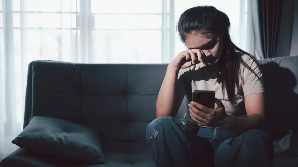 une jeunne femme sur un canapé sombre qui s'essuye l'oeil avec un téléphone à la main