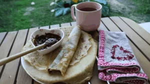 Table avec une assiette de crêpes, un tasse rose un serviette avec un coeur et de la Recette facile de la pâte à tartiner vegan choco-noisette dans un bol en forme de coeur