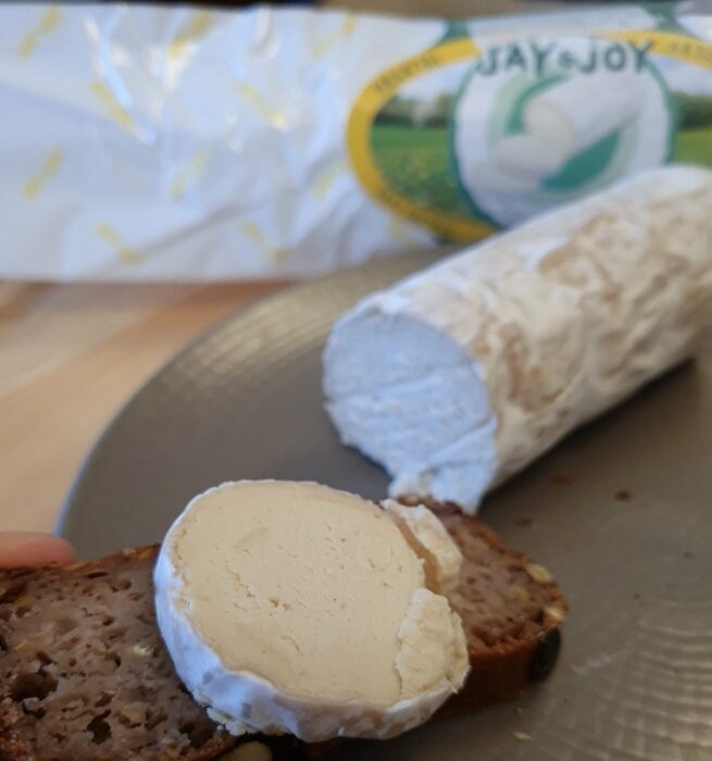 Bûche de chèvre vegan Jay & Joy Jil sur tranche de pain avec packaging dans le fond de la photo