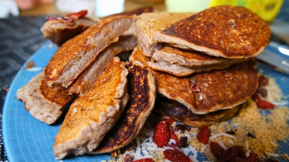 Pancake sans gluten coupé en deux sur une assiette bleu Degrenne avec garniture baies de goji, noix de coco râpée, poudre de noisette